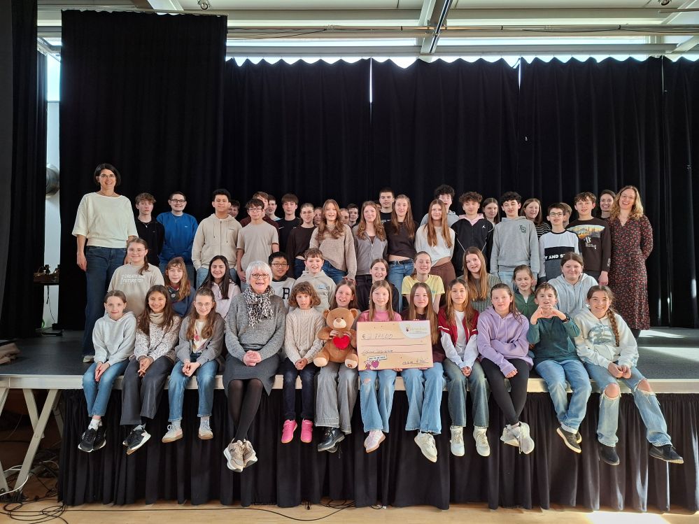 Schülerinnen und Schüler sowie die beteiligten Lehrkräfte überreichen in der Aula einen Scheck an eine Vertreterin von Bärenherz.