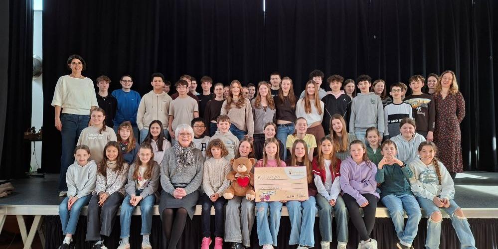 Schülerinnen und Schüler sowie die beteiligten Lehrkräfte überreichen in der Aula einen Scheck an eine Vertreterin von Bärenherz.