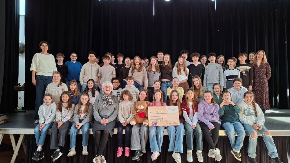Schülerinnen und Schüler sowie die beteiligten Lehrkräfte überreichen in der Aula einen Scheck an eine Vertreterin von Bärenherz.