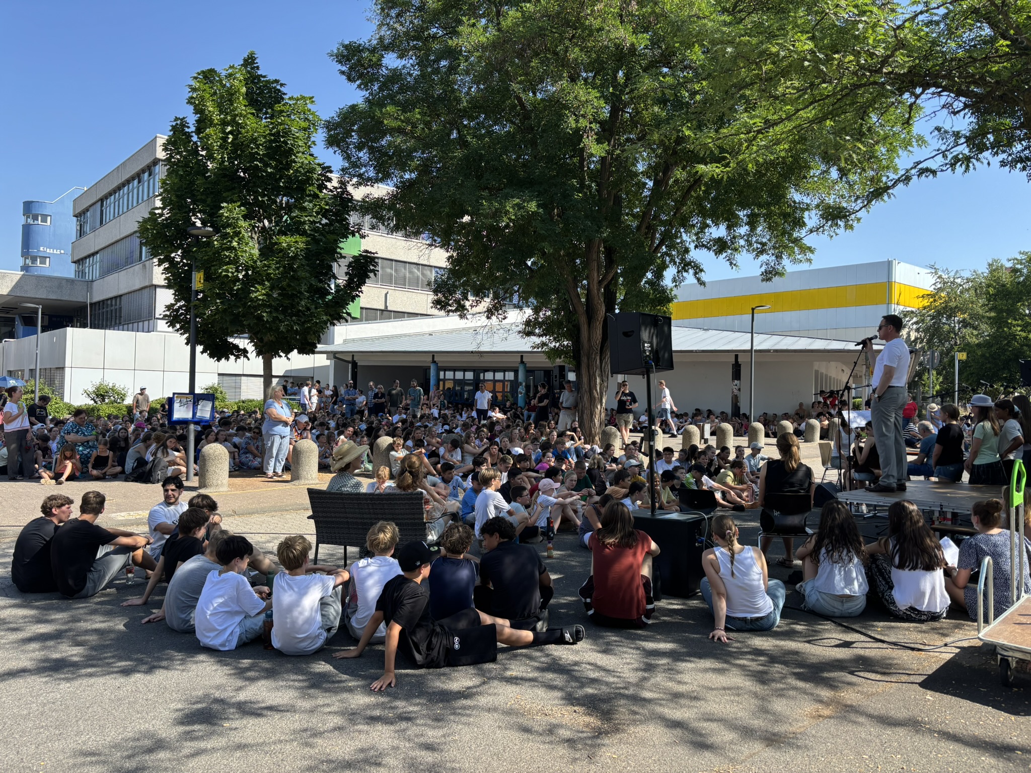 Kinder sitzen auf dem Schulhof, während Schulpfarrer Dr. Deutsch spricht.
